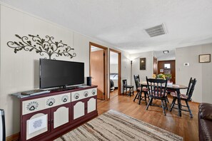 Una sala de estar con un televisor sobre un mueble, un comedor con sillas y una mesa, y una puerta que da a otra habitación.