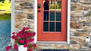 A stone building with a red door, a number '12' sign, a potted plant with pink flowers, and a decorative wreath.