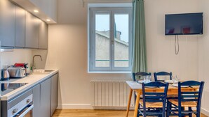 Eine moderne Küche mit Einbauofen, einem Essbereich mit Holztisch und blauen Stühlen, einem Fenster mit Blick auf Gebäude und einem an der Wand montierten Flachbildfernseher.