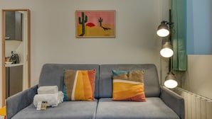 Ett modernt vardagsrum med en grå soffa, två färgglada kuddar, ett inramat konstverk som föreställer kaktusar och en vägglampa.