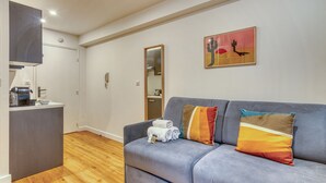 Ett modernt vardagsrum med en grå soffa, en liten köksdel och en tavla på väggen.