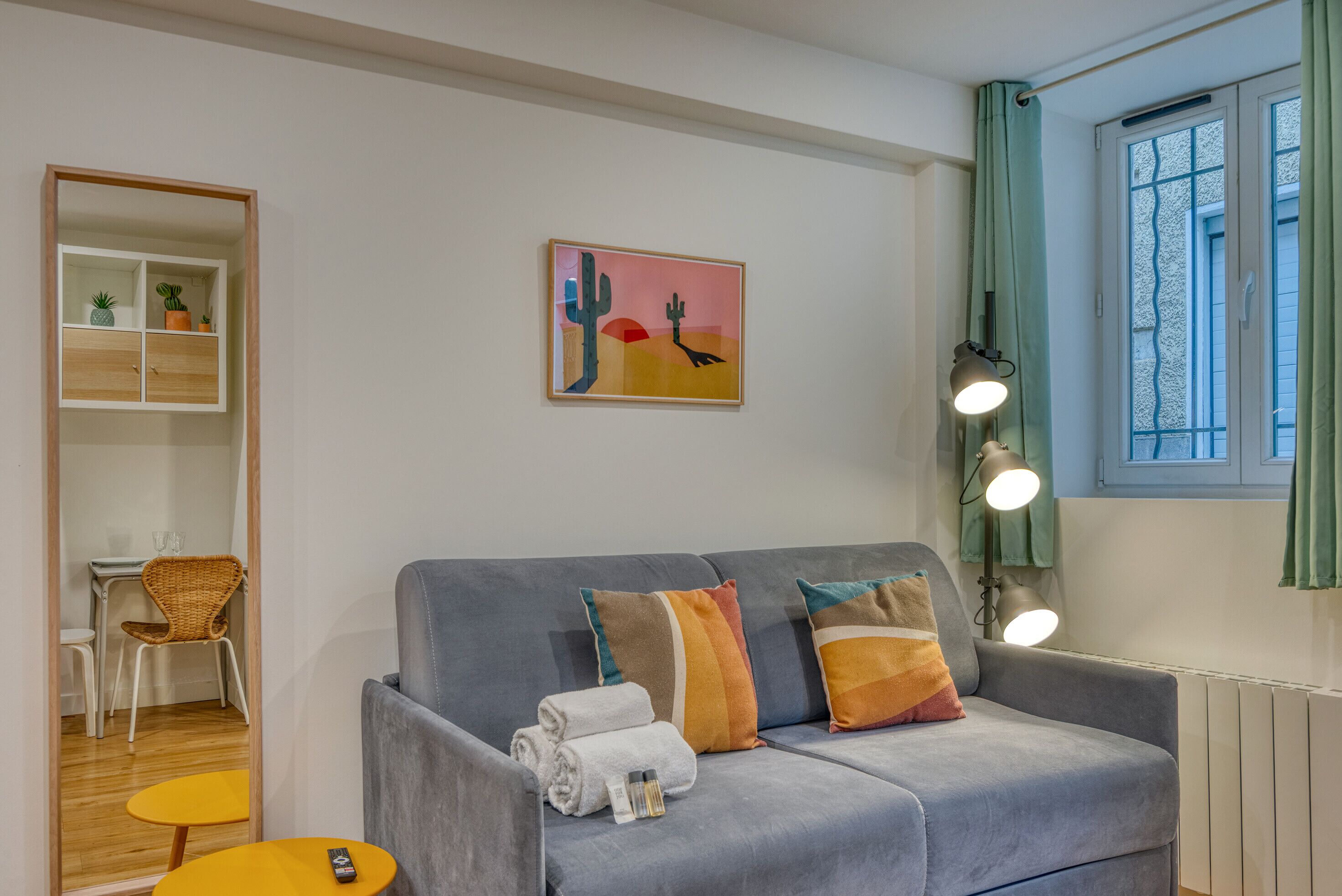Ein modernes Wohnzimmer mit einem grauen Sofa, einem kleinen gelben Tisch, einem Korbstuhl, einem an der Wand befestigten Regal mit Pflanzen und einem Gemälde einer Wüstenlandschaft.