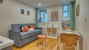 Ein gemütlicher Wohnbereich mit grauem Sofa, einem kleinen runden Tisch, einem Korbstuhl und einem weißen Schrank.