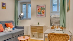 Ein gemütlicher Wohnraum mit Sofa, kleinem runden Tisch, Esstisch und Stühlen, Heizkörper und einem Fenster mit Vorhängen.