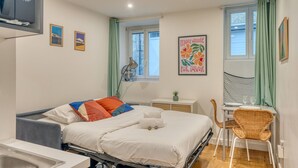 Ein Schlafzimmer mit einem Bett, einem Schreibtisch, einem Stuhl, einem Fenster mit Vorhängen, einem Wandventilator und einer kleinen Pflanze.