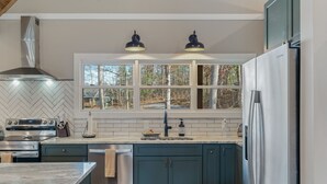 A modern kitchen with a herringbone tile backsplash, stainless steel appliances, and a window with a view of trees. - Mcdonald Lakeside Cottage (Sevierville)