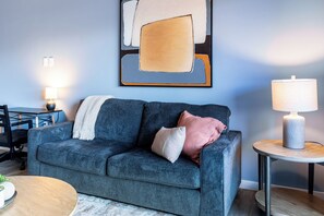 A modern living room with a dark blue sofa, a small round table with a lamp, and a framed abstract painting on the wall. - Landing Apartments Downtown Salt Lake City Near Trolley Square (Salt Lake City)