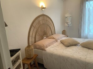 Une chambre avec une tête de lit en rotin, un lit recouvert d’une literie à motifs floraux, un petit tabouret en bois et une lampe fixée au mur.
