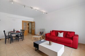 Un salon avec un canapé rouge, une table basse blanche, une table à manger avec des chaises et une porte en bois donnant sur une autre pièce.