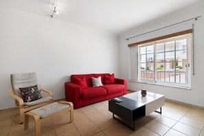 Un salon avec un canapé rouge, une chaise en bois, une table basse noire et une fenêtre donnant sur des immeubles.
