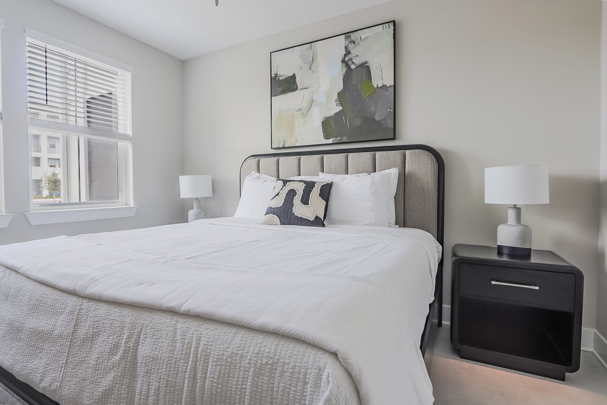 A modern bedroom with a large bed, two bedside tables with lamps, a window with blinds, and a piece of abstract art on the wall.