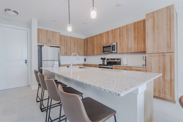 Une cuisine moderne avec des meubles en bois, un grand îlot central doté de tabourets, des appareils en acier inoxydable et un éclairage suspendu.