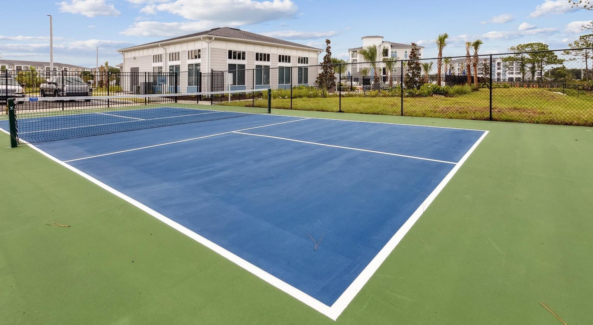 Un campo da tennis con una rete, circondato da una recinzione, con edifici sullo sfondo.