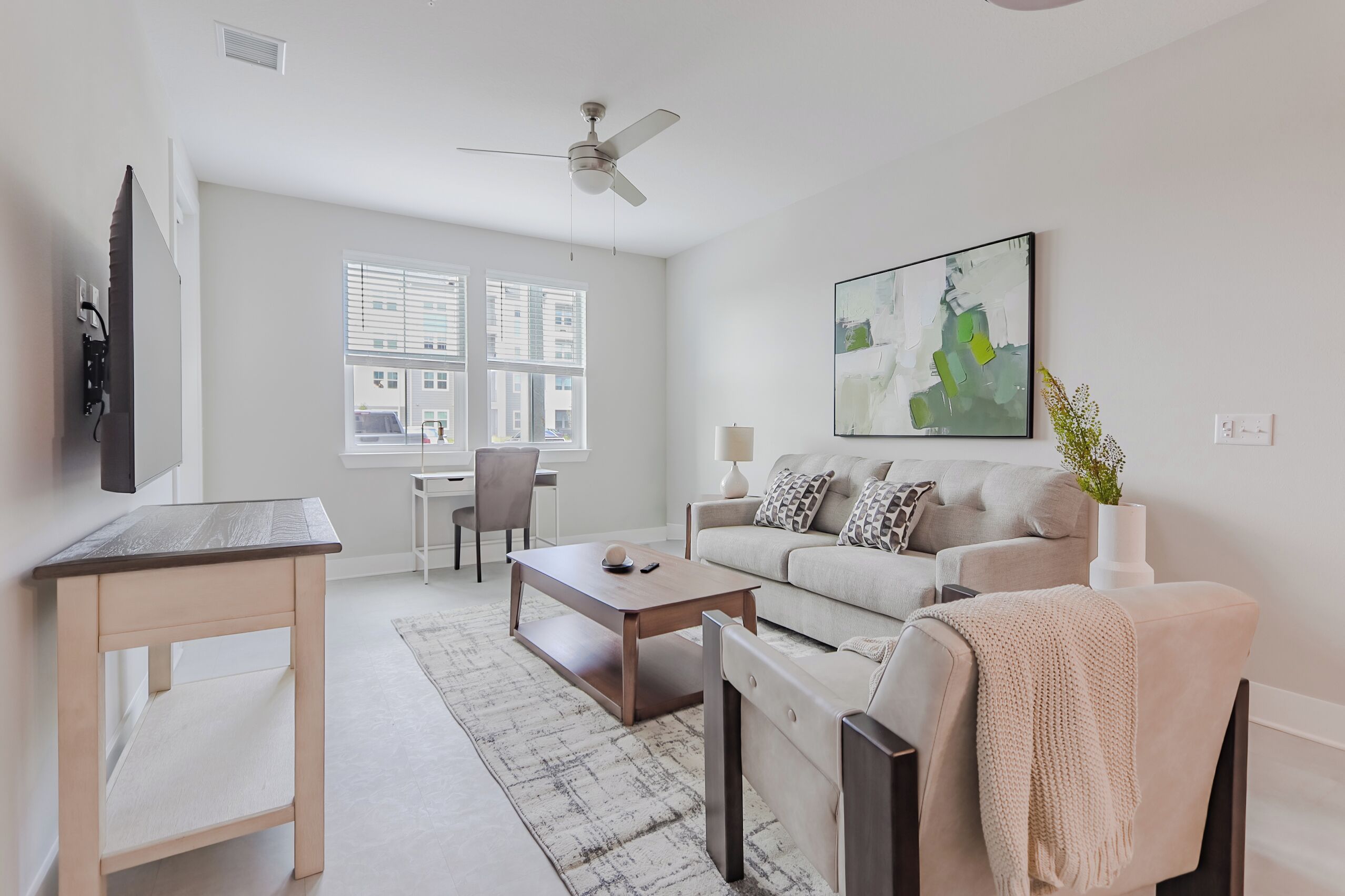 Un salon moderne avec un canapé, une table basse et un téléviseur. Il y a une fenêtre avec des stores, un ventilateur de plafond et un tableau au mur.
