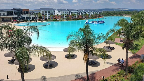 Ein Resort mit großem Schwimmbecken, Palmen und modernen Gebäuden im Hintergrund.