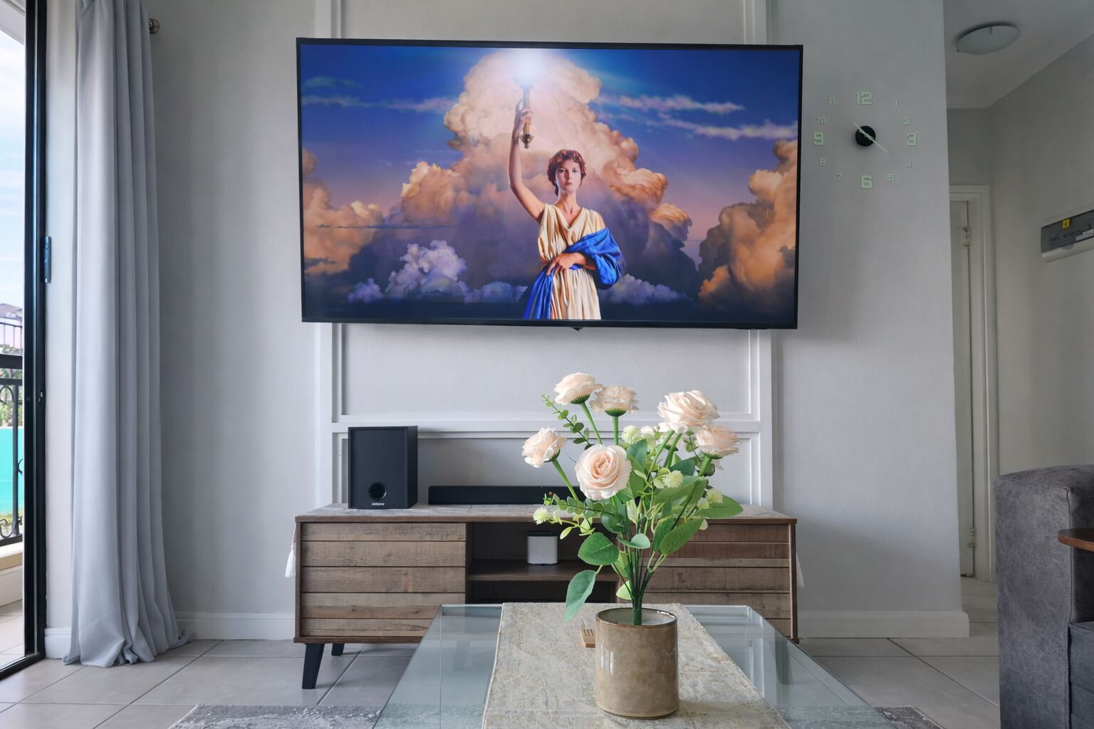 A living room with a flat-screen TV mounted on the wall, a wooden entertainment unit below, and a vase with flowers on a glass coffee table.