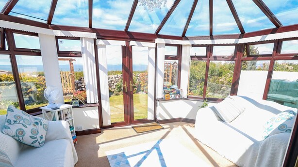 A sunroom with a glass ceiling, two white sofas, a small table with a fan, and a view of the garden and mountains. - Goetra (Harlech)