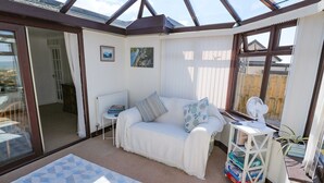 A sunlit room with a white sofa, a small table, and a radiator. There is a window with a view of a wooden fence and a building. - Goetra (Harlech)