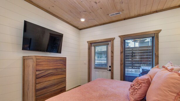 A bedroom with a wooden wardrobe, a television mounted on the wall, and a door with blinds. - Little Haven Chalet (Pigeon Forge)