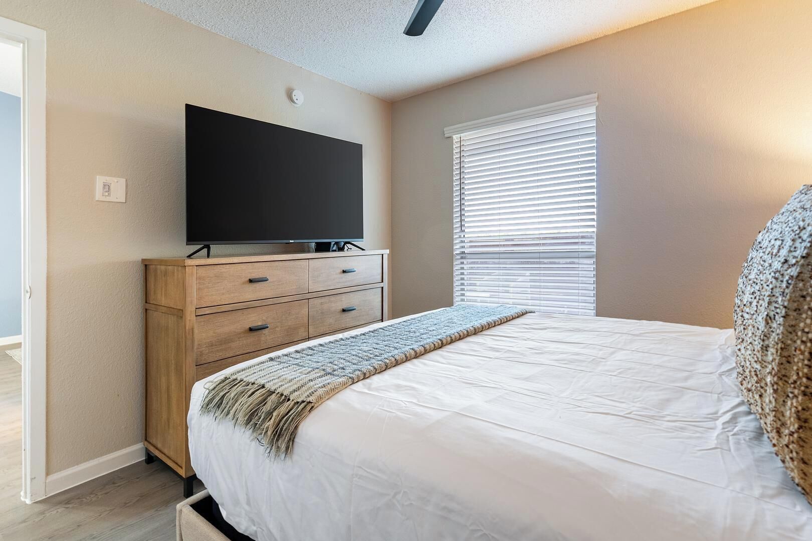 Une chambre à coucher avec un lit, un téléviseur posé sur une commode en bois et une fenêtre avec des stores.