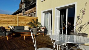 Un patio esterno con un pavimento in legno, un tavolo rotondo in metallo, sedie in metallo bianco, un divano con cuscini e una pianta in vaso.
