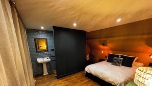 Una camera da letto con pavimento in legno, un letto bianco, un comodino verde, una parete nera, un lavandino bianco e uno specchio.