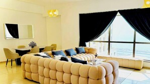 A modern living room with a large beige sectional sofa, a dining table with chairs, a small side table, and a large window with black curtains. - Premium Iconic Beachside JBR Stay (Dubai)