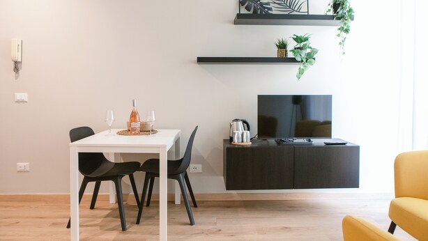 A modern living room with a white dining table, black chairs, a yellow armchair, a black entertainment unit with a TV, and a plant on a shelf. - [Bari Oberdan Home & Charm 1] Central Apartment (Bari)
