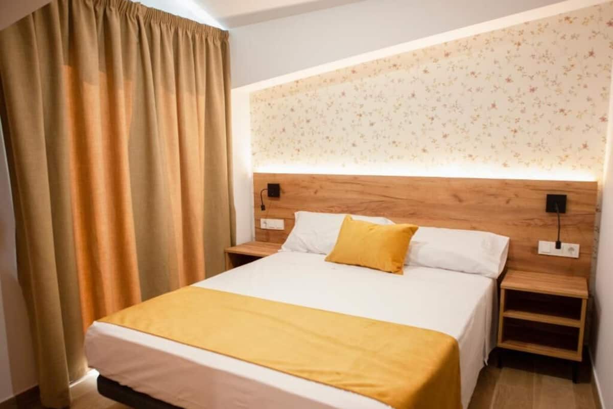 Una cama bien hecha con edredón blanco y amarillo, cabecera de madera y dos mesitas de noche con lámparas.