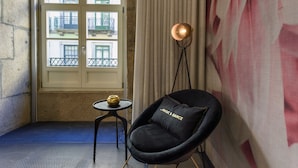 Una habitación acogedora con una silla negra, una mesita con un florero, una lámpara de pie y vistas a edificios a través de una ventana.
