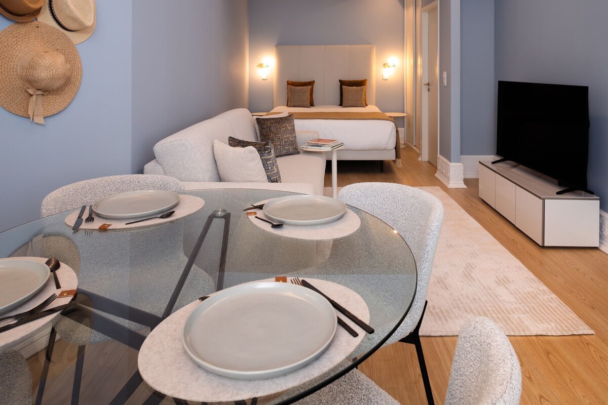 A modern hotel room with a glass dining table, a sofa, a TV, and a wall-mounted hat rack.
