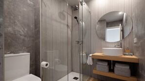 Une salle de bain moderne dotée d’une douche à parois de verre, de toilettes blanches, d’un meuble-lavabo en bois avec un miroir rond et d’une étagère intégrée où sont rangées les serviettes.