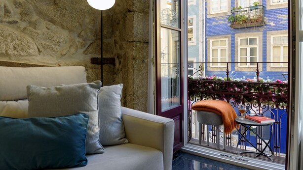A room with a stone wall, a sofa with cushions, a hanging light fixture, and a view of a blue building through a window. - RA - Baixa in Oporto (Porto)