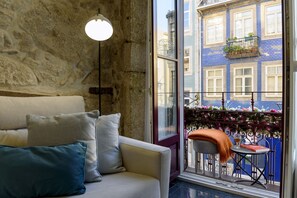 Una habitación con pared de piedra, un sofá con cojines, una lámpara colgante y vista a un edificio azul por la ventana.