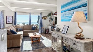A living room with a sofa, coffee table, and a lamp. There is a view of the ocean through the window. - Beachfront Aptos Home - Right on the Sand! (Aptos)