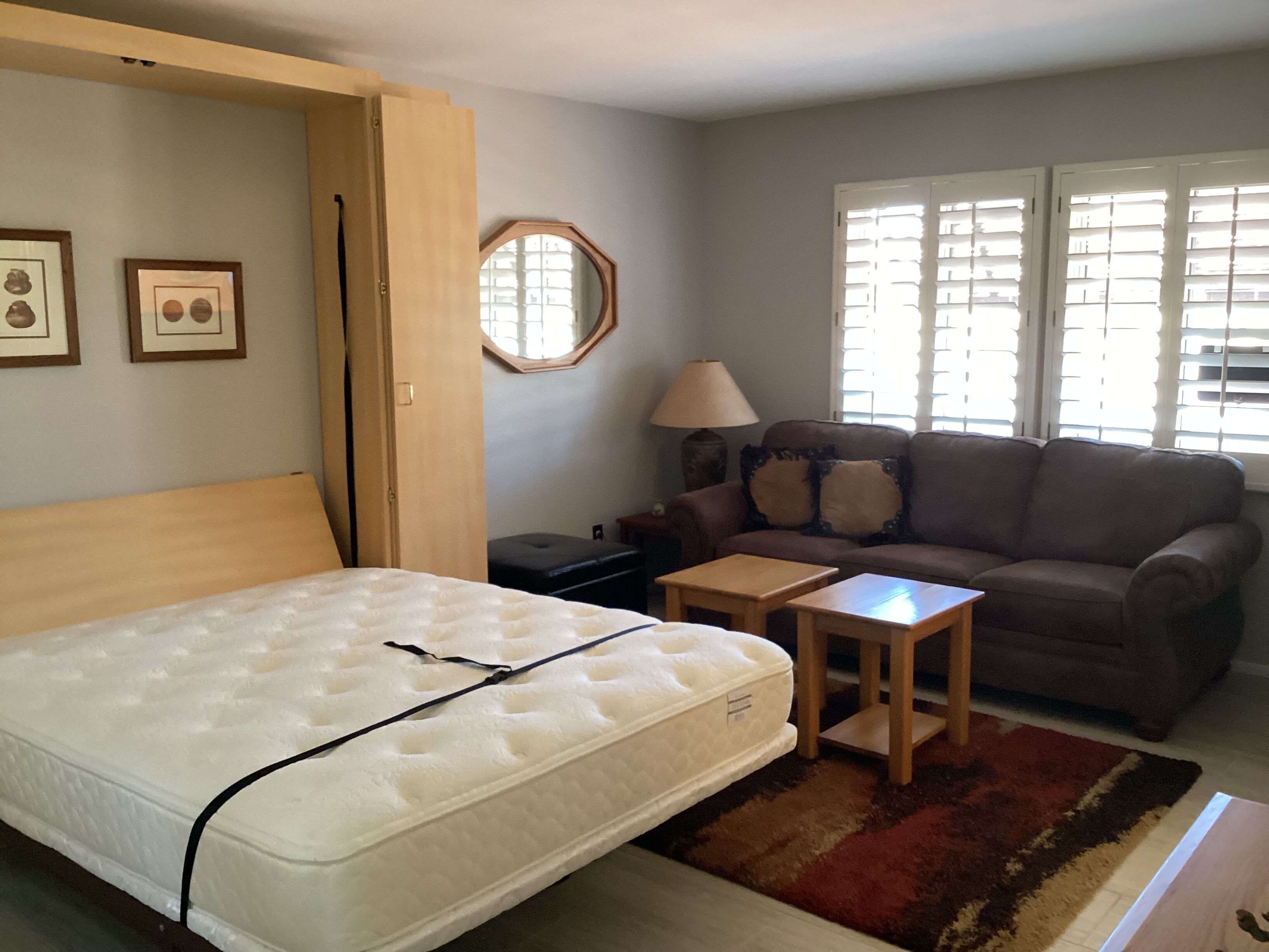 A bedroom with a bed, a wooden nightstand, a small table, a sofa, and a window with blinds.