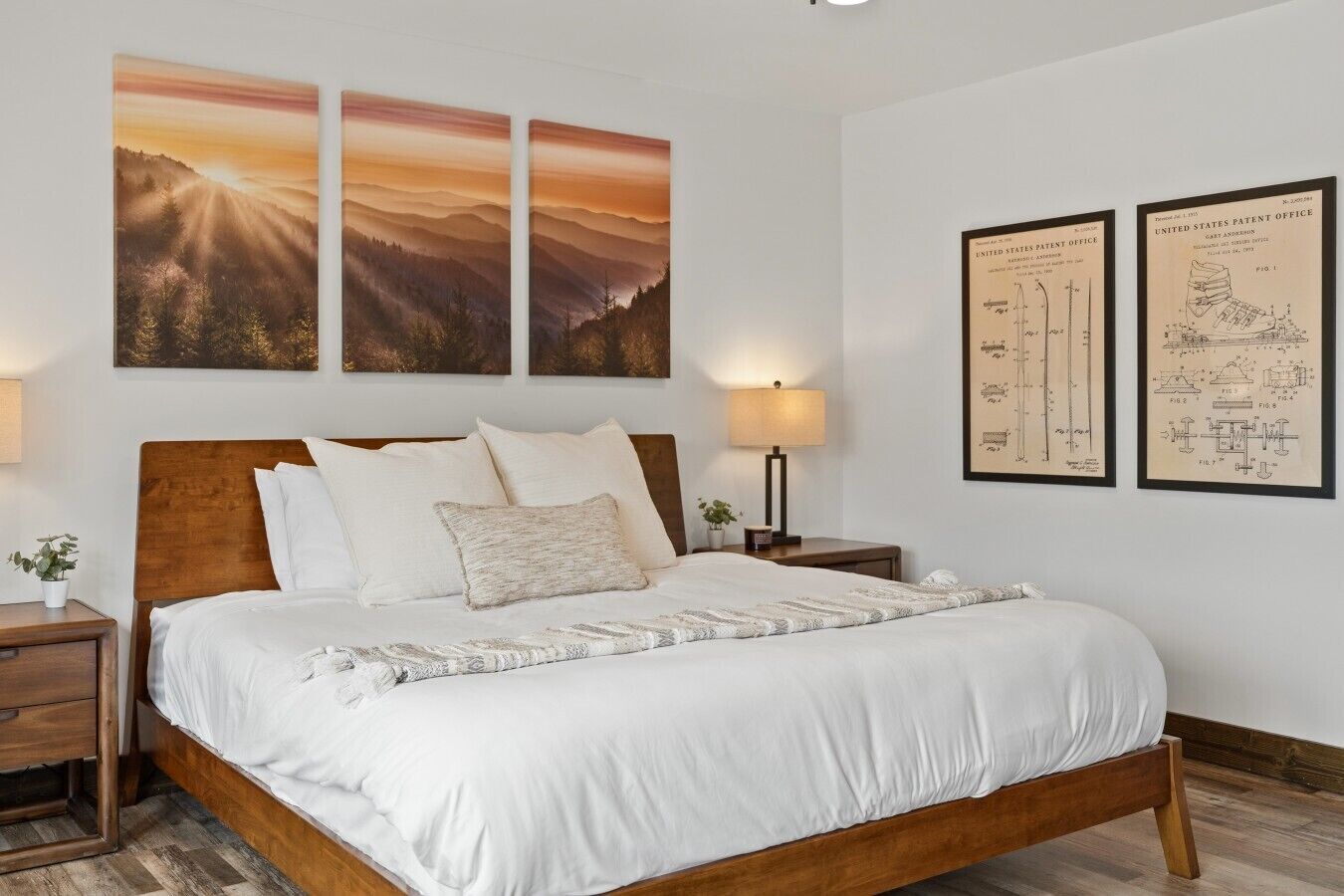A neatly made bed with a wooden headboard, two bedside tables with lamps, and three landscape paintings on the wall.