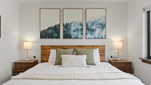 Un dormitorio con cabecera de madera, ropa de cama blanca, tres cuadros enmarcados sobre la cama y lámparas de noche sobre mesitas de madera.