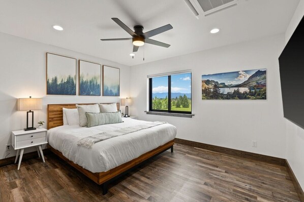 A bedroom with a bed, nightstands, a ceiling fan, and two windows with scenic views. - Peaks & Pines - Sweeping Mountain Views (Columbia Falls)