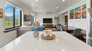 Una sala de estar moderna con un ventanal grande, un televisor de pantalla plana, una zona de comedor con mesa de madera y una cocina con refrigerador.
