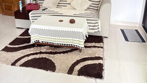 A room with a white sofa, a patterned rug, a small table with a white tablecloth, and a wall-mounted artwork.