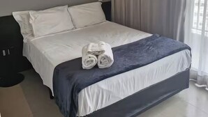 Una cama bien hecha con sábanas blancas y una toalla enrollada, un cuadro en la pared y una ventana con cortinas.