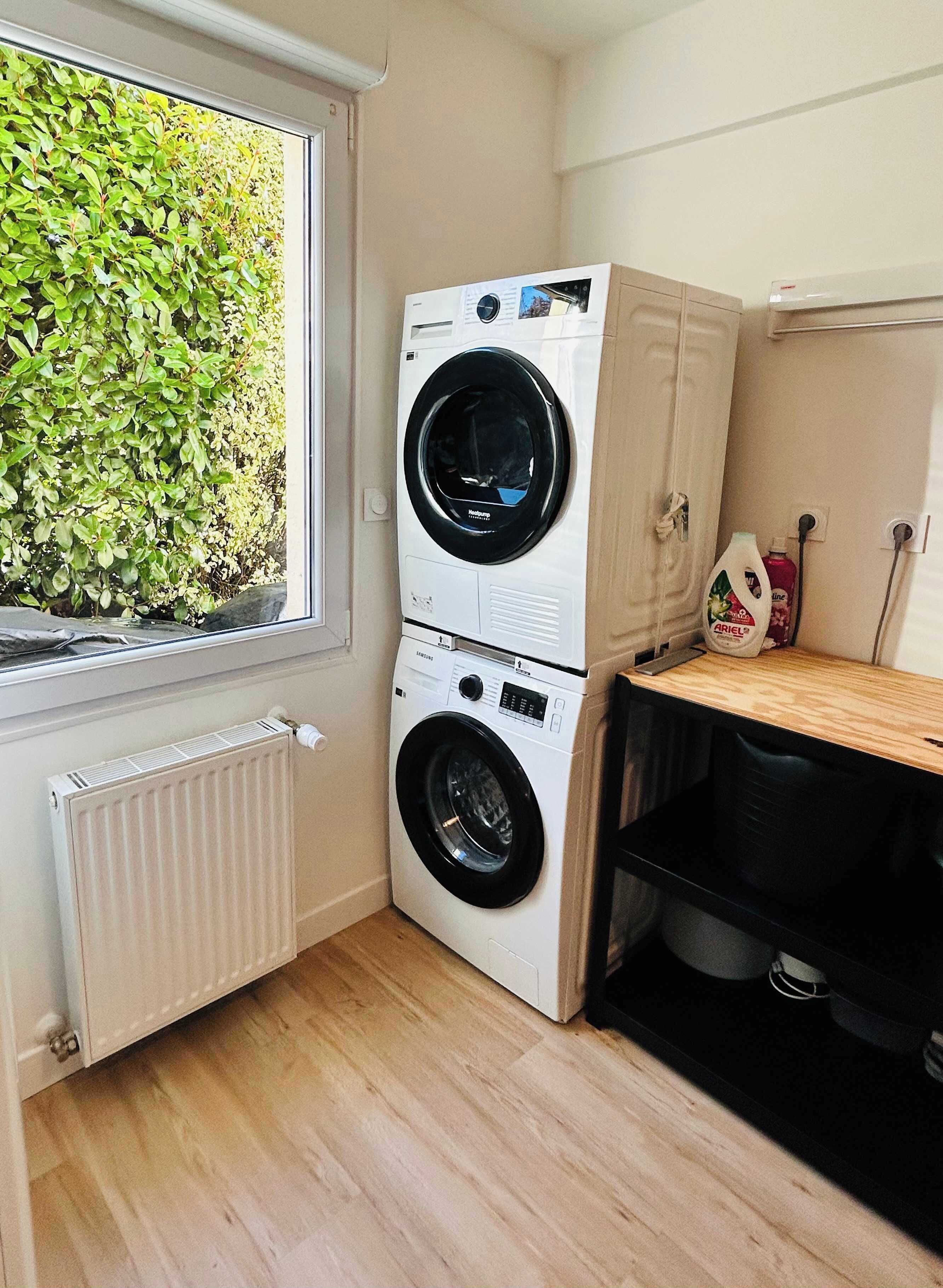 A laundry room with two stacked front-loading washing machines, a radiator, a window with a view of greenery, and a wooden shelf with a bottle.