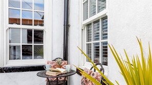 A white building with two windows, a small round table with a book and a cup, and a potted plant. - Boatman's Cottage Mousehole- Sleeps 4 -Garage (Mousehole)