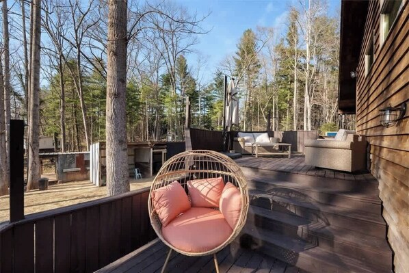 A wooden deck with a wicker chair and cushions, surrounded by trees. - Entire Farm for 23 | Hot Tub, Sauna, 15 Min to AVL (Asheville)