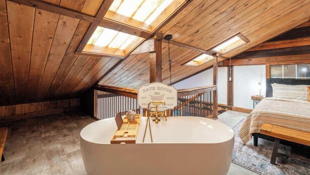 Un dormitorio acogedor en el ático con una bañera grande, vigas de madera y tragaluces.