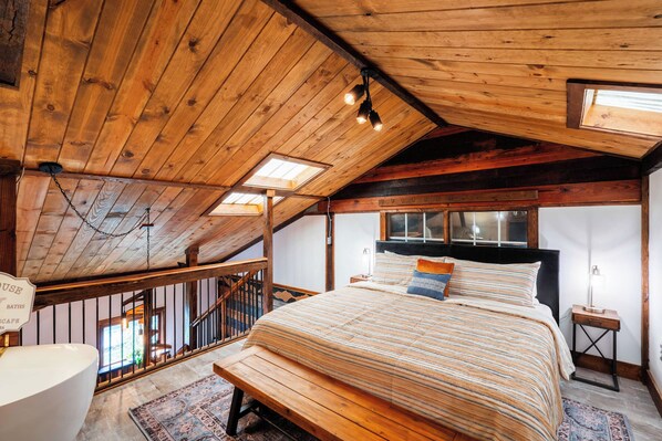 Un dormitorio con techo de madera, una cama con ropa de cama a rayas, un banco, una mesita de noche y un tragaluz.