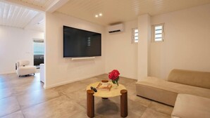 Ein modernes Wohnzimmer mit Flachbildfernseher an der Wand, einem beigefarbenen Sofa, einem runden Couchtisch mit einem Buch und einer Vase mit roten Blumen sowie einem weißen Sessel.