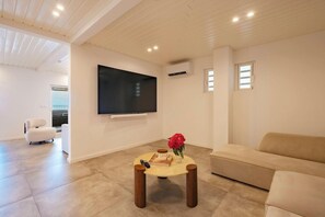 A modern living room with a flat-screen TV mounted on the wall, a beige sofa, a round coffee table with a book and a vase of red flowers, and a white armchair. - Amazing ocean front 6-bedroom villa in Grand-Case Saint-Martin (Grand-Case)