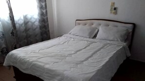 Ein ordentlich bezogenes Bett mit weißer Bettwäsche in einem Zimmer mit gefliestem Boden und einem Vorhang mit geometrischem Muster.